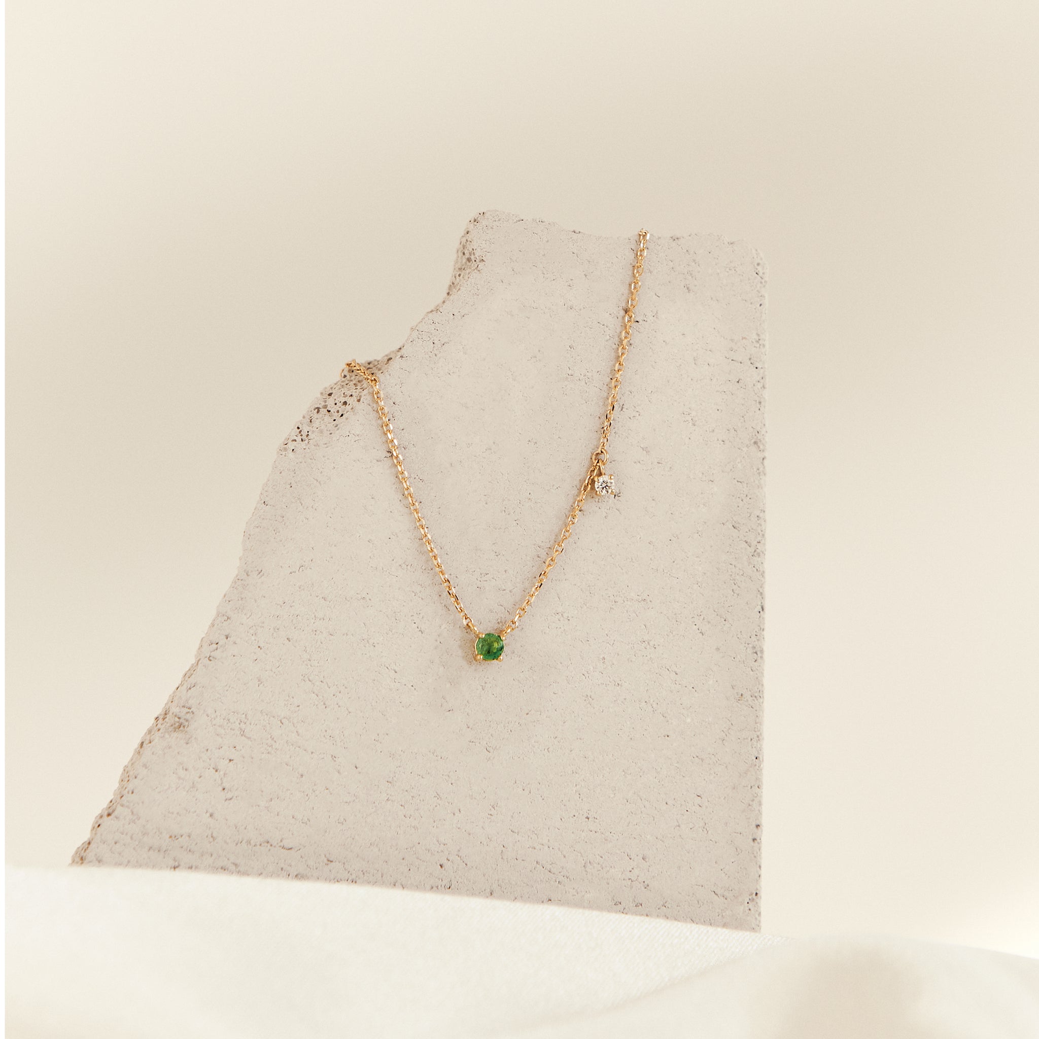 A gold necklace with a small green gemstone pendant fixed on the center of the chain with an additional clear side stone dangling off the chain about 2cm to the right