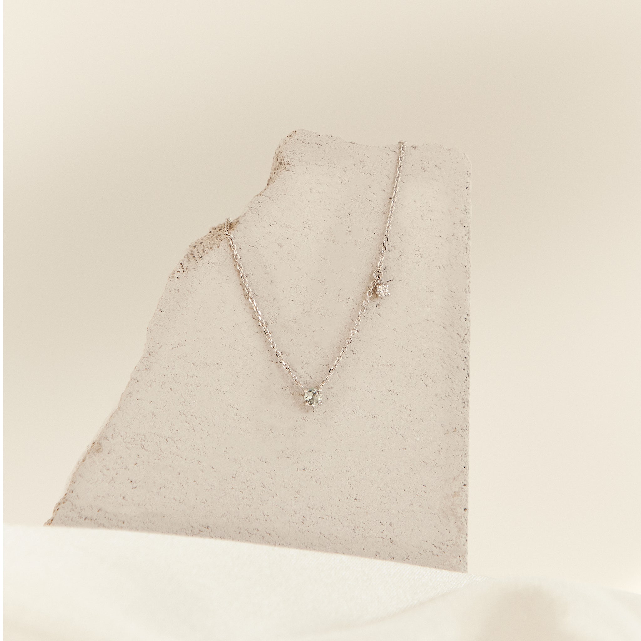 A white gold necklace with a small clear gemstone pendant fixed on the center of the chain with an additional clear side stone dangling off the chain about 2cm to the right