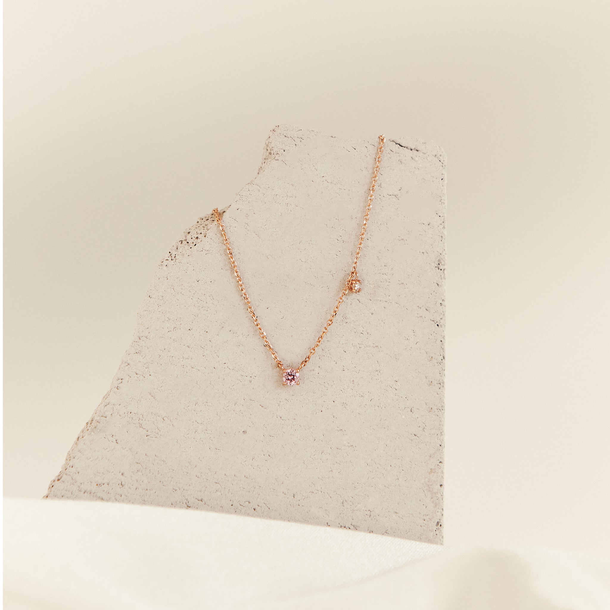 A rose gold necklace with a small pink gemstone pendant fixed on the center of the chain with an additional clear side stone dangling off the chain about 2cm to the right